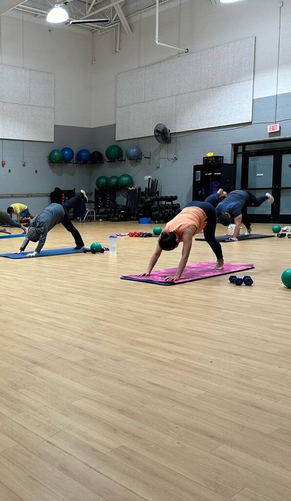 Group fitness class participants performing yoga poses on mats in a spacious gym with exercise equipment in the background.