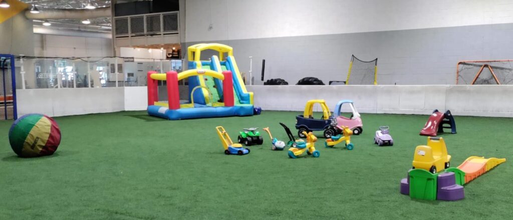 A bounce house, bouncy slide, and several toy trucks and bikes set up on a turf field inside