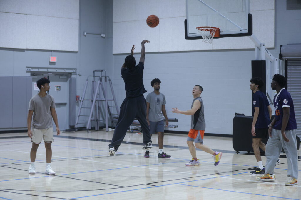 A boy in a navy blue shirt and sweats shoots a basketball while several other players watch